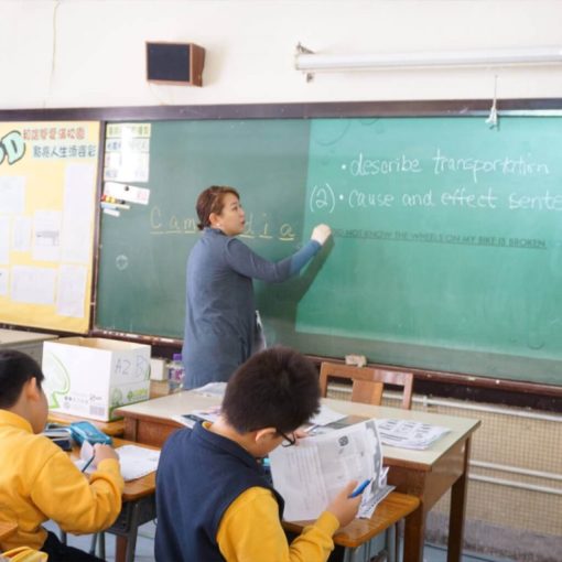 Female English teacher writing on a blackboard in a classroom with Hong Kong primary school students.